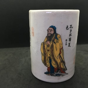 Könnte beinhalten: Zylindrische Tasse aus weißer Keramik mit einer gemalten Illustration eines Mannes in gelben Gewändern. Die Tasse hat chinesische Schriftzeichen und einen roten Stempel. Der Mann hat einen langen Bart und trägt einen blauen Hut. Die Tasse hat ein Craquelé-Finish.