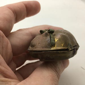 May include: A small, antique, bronze-colored trinket box with a hinged lid. The box has a rounded shape and a small green bow on top. The edges of the box show hints of gold. The box is held in a person's hand.