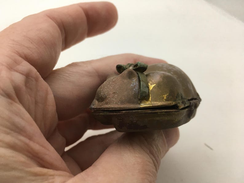 May include: A small, antique, bronze-colored trinket box with a hinged lid. The box has a rounded shape and a small green bow on top. The edges of the box show hints of gold. The box is held in a person's hand.