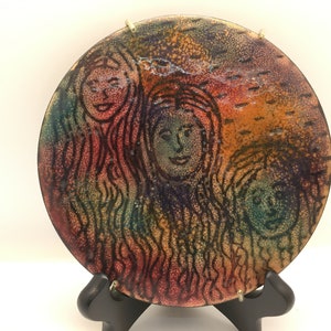 May include: A round ceramic plate with a colorful, abstract design featuring three faces. The faces are outlined in black and have a speckled, textured background. The plate is on a black stand.