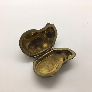 May include: An open, antique brass-colored trinket box. The box is shaped like a pear and has a hinged lid. The interior of the box is visible, showing a tarnished, aged appearance. The box is on a white surface.