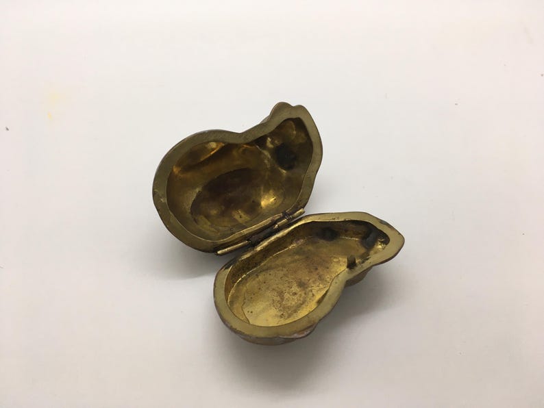 May include: An open, antique brass-colored trinket box. The box is shaped like a pear and has a hinged lid. The interior of the box is visible, showing a tarnished, aged appearance. The box is on a white surface.