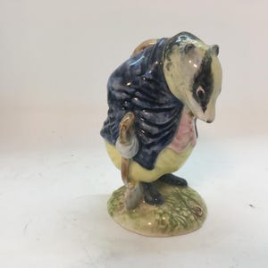 May include: A ceramic figurine of a badger dressed in a blue coat, pink vest, and yellow trousers, holding a small shovel. The badger stands on a green grassy base, with a black and white head.