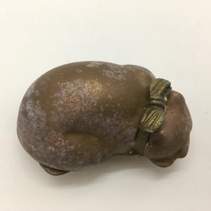 May include: A bronze-colored cat figurine with a bow around its neck. The cat is in a curled-up position, with a weathered appearance. The bow is a darker shade of bronze, adding a contrasting detail to the sculpture.