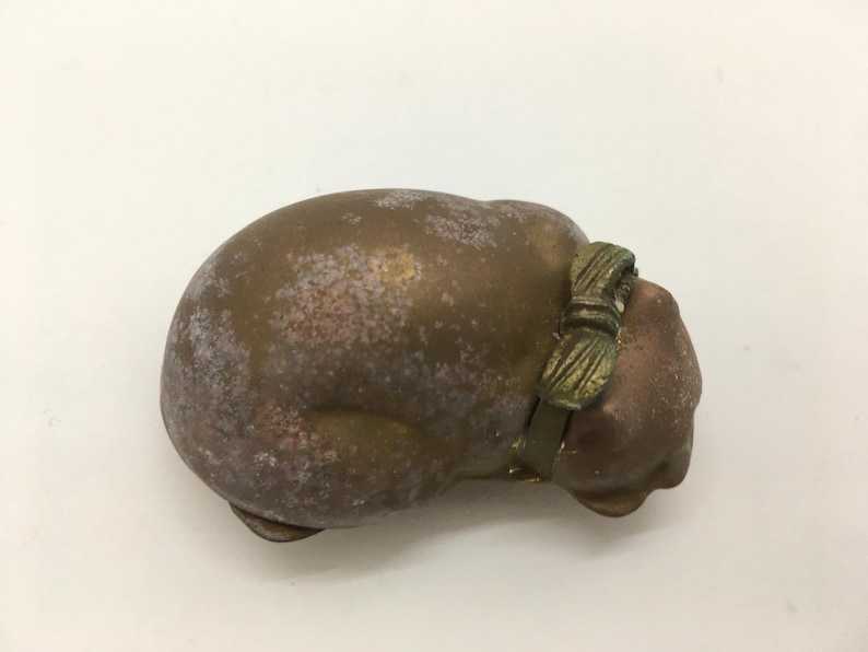 May include: A bronze-colored cat figurine with a bow around its neck. The cat is in a curled-up position, with a weathered appearance. The bow is a darker shade of bronze, adding a contrasting detail to the sculpture.