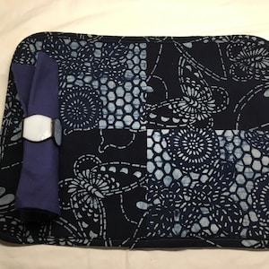 May include: A navy blue placemat with a white and blue floral pattern. A rolled-up purple napkin is held by a white and silver napkin ring. The placemat is rectangular with rounded corners.