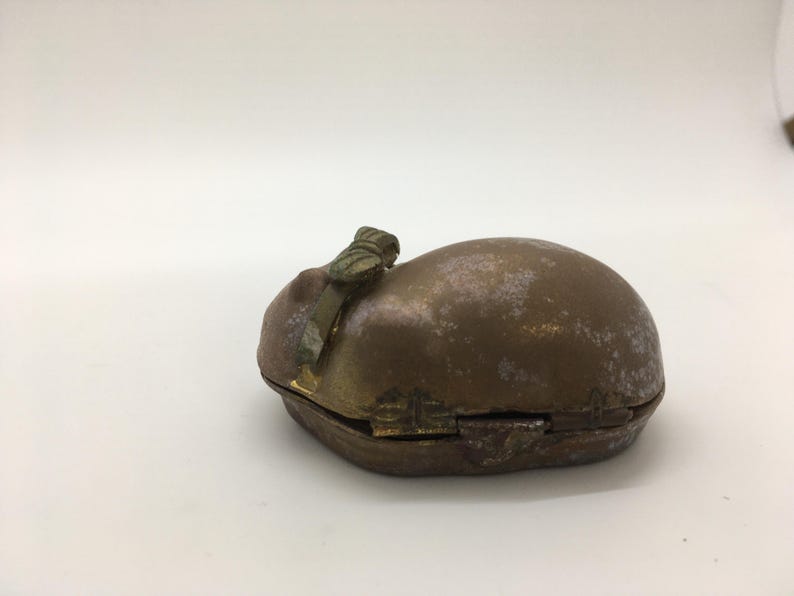 May include: A small, antique brass box with a hinged lid. The box has a rounded, oval shape and a tarnished, aged appearance. A decorative clasp and handle are visible on the top. The box is likely used for storing small items.