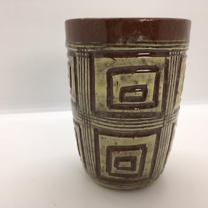 May include: A brown and beige ceramic tumbler with a geometric square pattern. The top rim is a darker brown. The tumbler is cylindrical and appears to be handmade.