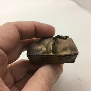May include: A small, antique metal cat-shaped trinket box. The box is a muted gold color with a darker, tarnished finish. The cat is depicted curled up, with a small clasp on top. The box is held in a hand.