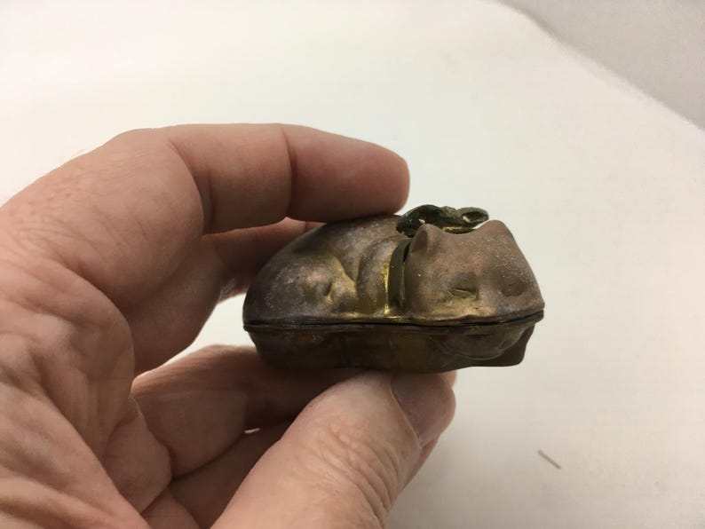 May include: A small, antique metal cat-shaped trinket box. The box is a muted gold color with a darker, tarnished finish. The cat is depicted curled up, with a small clasp on top. The box is held in a hand.