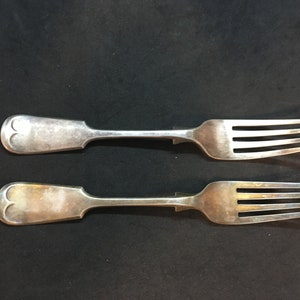 Puede incluir: Dos tenedores de plata con mangos ornamentados. Los tenedores están dispuestos uno al lado del otro sobre una superficie negra.