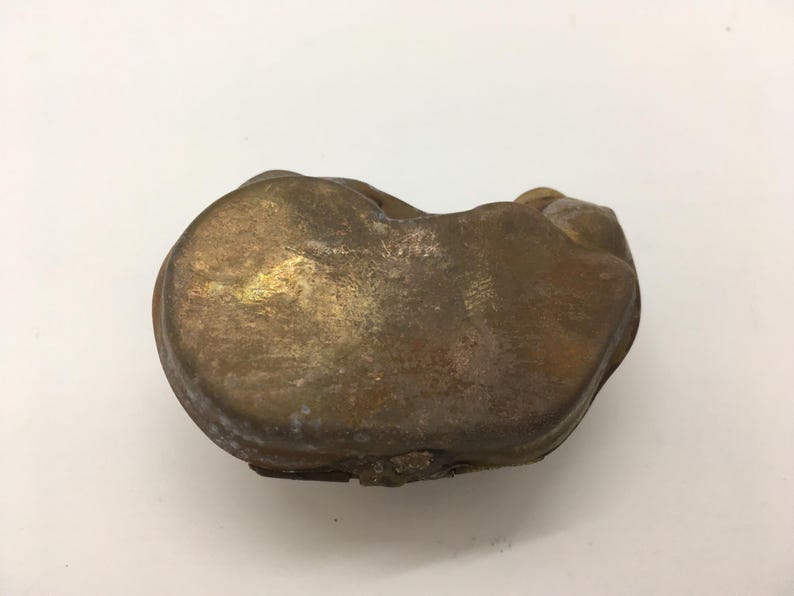 May include: A small, antique brass box with a rounded, organic shape. The box has a tarnished, aged appearance with variations in color from gold to brown. The lid is slightly raised, suggesting it may open. The object is set against a white background.