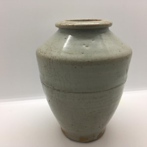 May include: A light gray ceramic vase with a wide, rounded body and a narrow, thick-rimmed opening. The surface features a visible crackle glaze pattern throughout, with subtle variations in tone and a smooth, glossy finish on the exterior.
