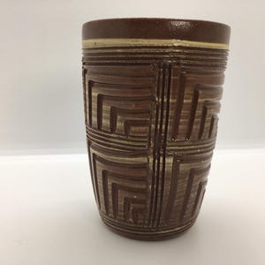 May include: A brown ceramic tumbler with a geometric pattern. The tumbler has a cylindrical shape with a slightly flared top. The design features a repeating pattern of squares and lines in a lighter brown color. The rim has a thin, light-colored band.
