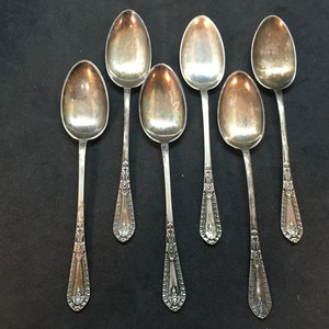 May include: Six silver spoons with ornate designs on the handles. The spoons are arranged in a row on a black surface.