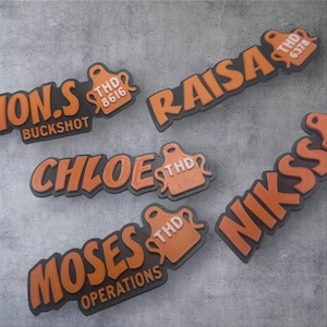 May include: Several orange and black name tags with various names and text, including "RAISA", "CHLOE", "MOSES OPERATIONS", and "NIKSS". Each tag has a small orange tag with "THD" and a number. The tags are on a gray surface.