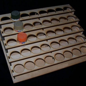 May include: A wooden paint rack with five tiers, each tier has 12 circular slots for holding paint bottles. The rack is made of light brown wood and has a natural finish. There are three paint bottles in the slots, one green, one orange, and one beige.