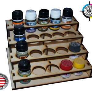May include: Wooden paint bottle storage rack with five shelves and 24 round holes for holding small bottles. The rack is made of light brown wood and has a white label with the text "MADE IN THE USA" on the front. The rack is also labeled with "DONALD ETHREDGE ENTERPRISES" on the top shelf.