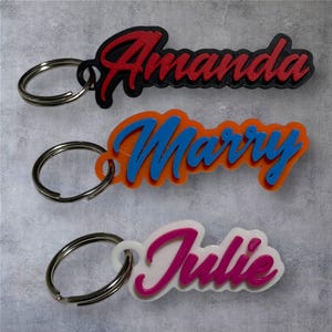 May include: Three personalized keychains with names: "Amanda" in red on black, "Marry" in blue on orange, and "Julie" in pink on white. Each keychain has a silver metal ring for attaching to keys or bags. The names are in a cursive font.