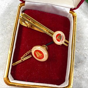 May include: A gold tie tack and tie clip set in a red velvet box. Both pieces feature the Northeast Normal University logo, which includes a sailboat and the text "Northeast Normal University Changchun China".