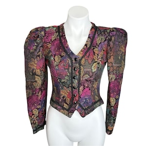May include: A floral print jacket with a V-neck and black buttons. The jacket features a colorful pattern of roses and leaves in shades of pink, purple, and gold. Black trim accents the edges and button line. The jacket has long sleeves and a fitted silhouette.