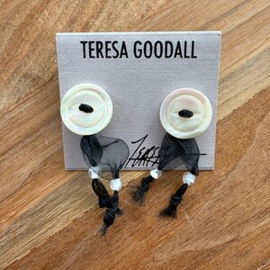 May include: Earrings with white button-like tops and black mesh and beaded accents. The earrings are displayed on a gray card with the text "TERESA GOODALL". The earrings have a unique, handcrafted appearance.