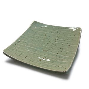 May include: A square, ceramic plate with a light teal glaze and a textured, ridged surface. The plate has small, dark brown speckles throughout and rounded corners. The plate is slightly elevated, casting a soft shadow.
