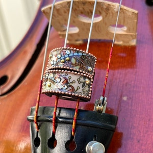 Könnte beinhalten: Nahaufnahme eines dekorativen Rings auf den Saiten einer Geige. Der Ring ist mit kleinen, farbigen Edelsteinen und einem floralen Muster verziert. Steg und Korpus der Geige sind im Hintergrund zu sehen.