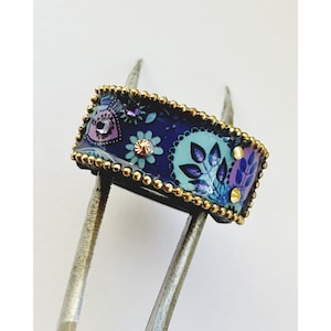 May include: A wide, gold-rimmed ring with a blue, purple, and green floral pattern. The ring is decorated with small gold beads and rhinestones.