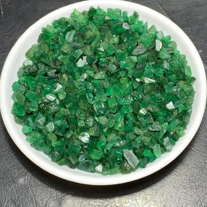 Può includere: Una ciotola bianca piena di numerose piccole gemme di smeraldo grezze. Gli smeraldi sono di un colore verde intenso e vibrante, con forme e dimensioni variabili. La ciotola è rotonda e poggia su una superficie scura.