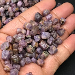 Puede incluir: Un puñado de piedras preciosas de rubí en bruto, sin cortar, en varios tonos de púrpura y marrón. Las piedras rugosas tienen un aspecto natural, sin pulir, con estructuras cristalinas y texturas visibles. Las piedras miden aproximadamente 0,6 a 1,9 cm.