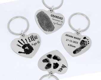 May include: Four silver heart-shaped keychains with black ink imprints of fingerprints, handprints, and footprints. The keychains are personalized with names, dates, and messages like "Grandad Forever in my heart", "Ellie Age 6", "Ariana Marie 27-12-2023 6:05am", and "Max".