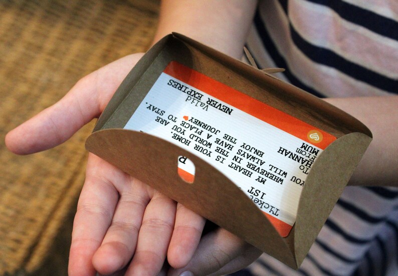 Pu&ograve; includere: Una busta di carta marrone con un biglietto all'interno. Il biglietto &egrave; arancione e bianco con il testo "Ticket 1st", "My heart is your home. Wherever in the world you are, enjoy the journey. You will always have a place to stay." e "To Hannah From Mum ♡ Valid Never Expires".