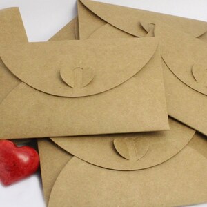 Pu&ograve; includere: Un mucchio di buste di carta marrone con un ritaglio a forma di cuore sul lembo. Una pietra rossa a forma di cuore si trova davanti alle buste.