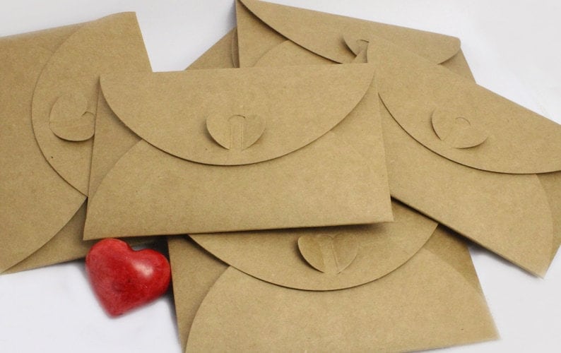 Pu&ograve; includere: Un mucchio di buste di carta marrone con un ritaglio a forma di cuore sul lembo. Una pietra rossa a forma di cuore si trova davanti alle buste.
