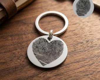 May include: A silver keychain with a circular pendant featuring a fingerprint in the shape of a heart. The keychain is on a wooden surface, with a key and a small notebook in the background.