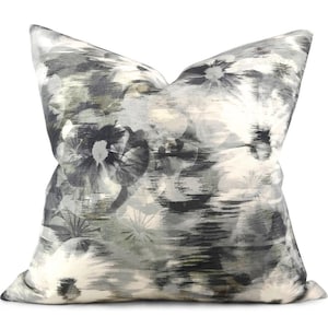 May include: A square throw pillow featuring an abstract floral pattern in shades of charcoal gray, slate, white, and muted olive green. The design displays blurred, painterly flower shapes with dark centers and soft, streaked petal edges.