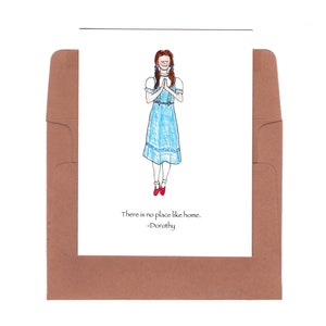 Könnte beinhalten: Eine Grußkarte mit einer handgezeichneten Illustration von Dorothy aus dem Zauberer von Oz. Dorothy trägt ein blaues Gingham-Kleid und rote Schuhe. Die Karte trägt den Spruch "Es gibt keinen Ort wie zu Hause. -Dorothy" aufgedruckt.