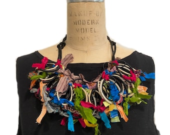 Puede incluir: Un collar colorido hecho de retazos de tela y gomas elásticas. El collar se muestra en un maniquí con una camisa negra.