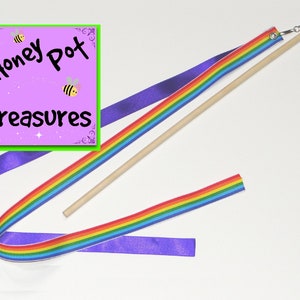 May include: Rainbow ribbon wand with a wooden stick and a bell on the end. The ribbon is attached to the stick with a metal ring. The wand is on a white background. The image also includes a pink square with the text "Honey Pot Treasures".