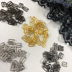 May include: A close-up of a variety of bra strap sliders in gold, silver, and black metal. The sliders are arranged in piles on a white surface.