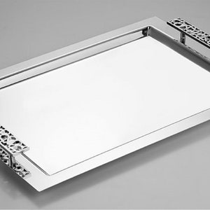 Puede incluir: Una bandeja de servir rectangular de plata con asas decorativas recortadas. La bandeja es rectangular y tiene una superficie pulida y reflectante.