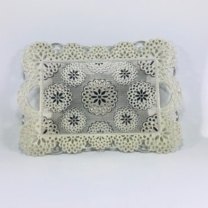 Op de afbeelding: Een witte serveerschaal met een decoratief zilveren bloemenpatroon. De schaal heeft een gegolfde rand en twee handvatten.