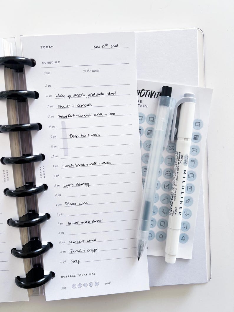 Op de afbeelding: Een planner met een dagschema, inclusief tijden en activiteiten zoals 'Wake up, stretch, gratitude ritual' en 'Pilates class'. Een doorzichtige pen en een witte markeerstift liggen naast de planner, samen met een stickervel. De datum 'Nov 12th, 2025' staat bovenaan.