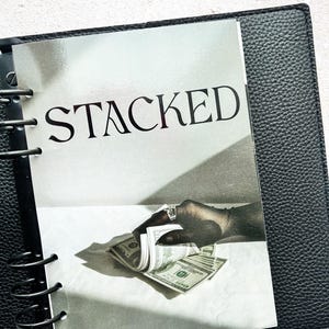 Pu&ograve; includere: Un raccoglitore in pelle nera con un inserto stampato con la parola "STACKED" in lettere grandi e in grassetto. L'inserto mostra una mano con un anello che tiene in mano una pila di banconote da cento dollari USA. Il raccoglitore &egrave; aperto.