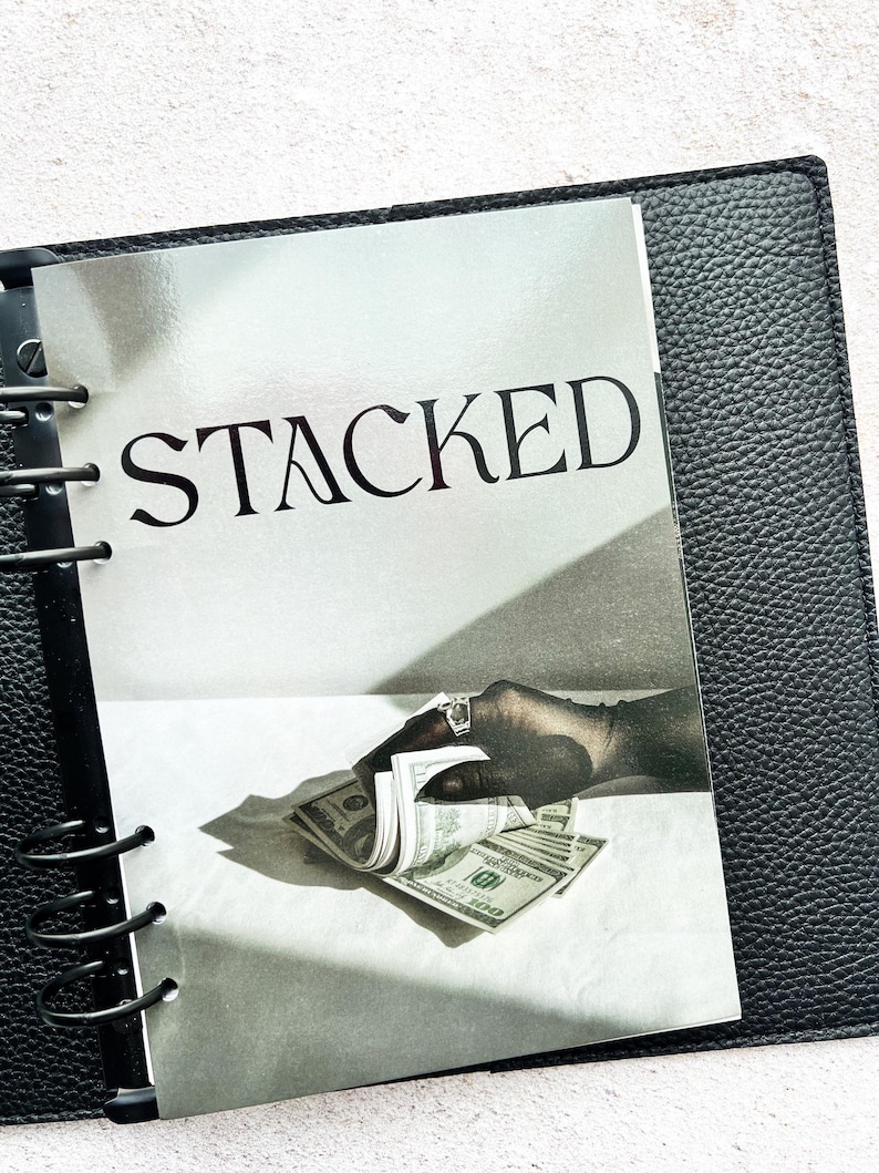 Pu&ograve; includere: Un raccoglitore in pelle nera con un inserto stampato con la parola "STACKED" in lettere grandi e in grassetto. L'inserto mostra una mano con un anello che tiene in mano una pila di banconote da cento dollari USA. Il raccoglitore &egrave; aperto.