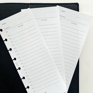 Op de afbeelding: Witte plannerpagina's met een schema-indeling, met de woorden "TODAY" en "SCHEDULE". De pagina's hebben voorgedrukte tijdvakken en lijnen voor notities, met de zin "On the agenda" bovenaan. De pagina's liggen op een zwart, leerachtig oppervlak.
