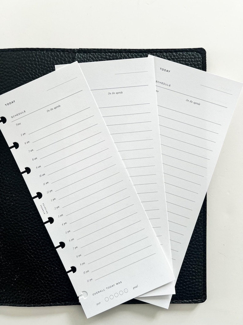 Op de afbeelding: Witte plannerpagina's met een schema-indeling, met de woorden "TODAY" en "SCHEDULE". De pagina's hebben voorgedrukte tijdvakken en lijnen voor notities, met de zin "On the agenda" bovenaan. De pagina's liggen op een zwart, leerachtig oppervlak.