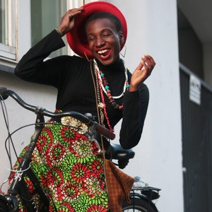 May include: A black bicycle with a woman wearing a black turtleneck and a red hat. She is wearing a colourful skirt with a floral pattern in red, green, and white. The woman is smiling and wearing multiple necklaces.