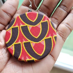 May include: A pair of large, round earrings with a geometric pattern in red, black, and yellow. The earrings are made of fabric and have a silver hook.